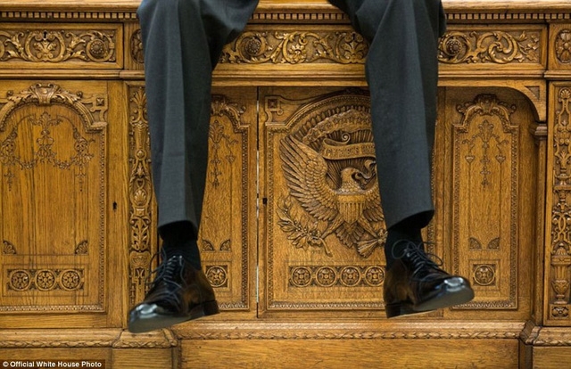 Tổng thống Obama ngồi trên bàn làm việc trong khi đang nói chuyện với hai phụ tá trong Phòng bầu dục.